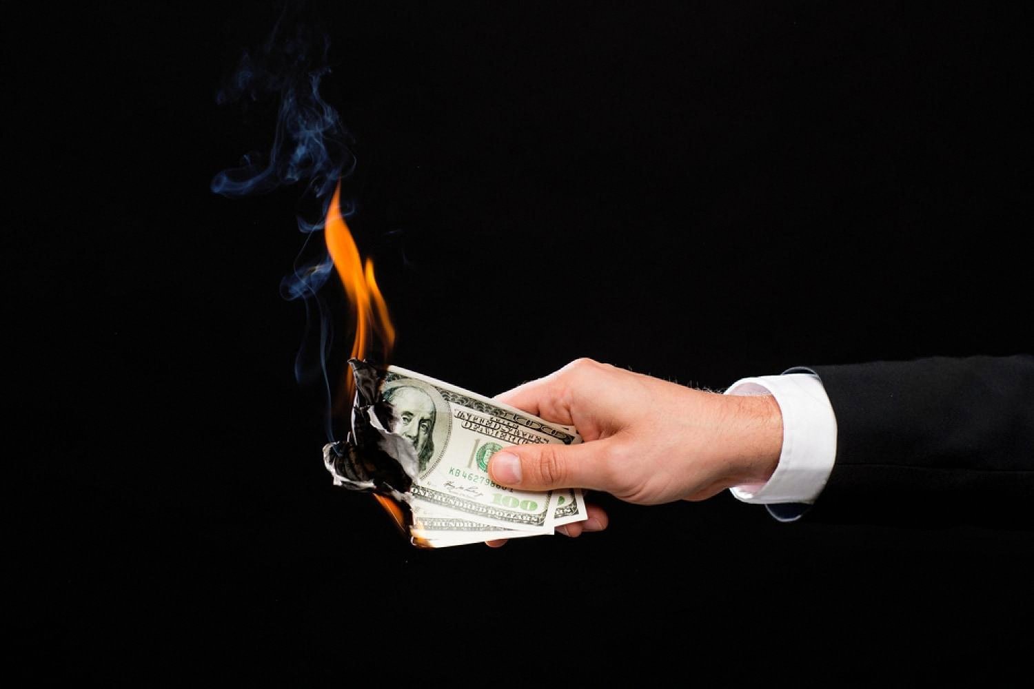 Hand holding burning US dollar bills against a black background, symbolising wasted wealth